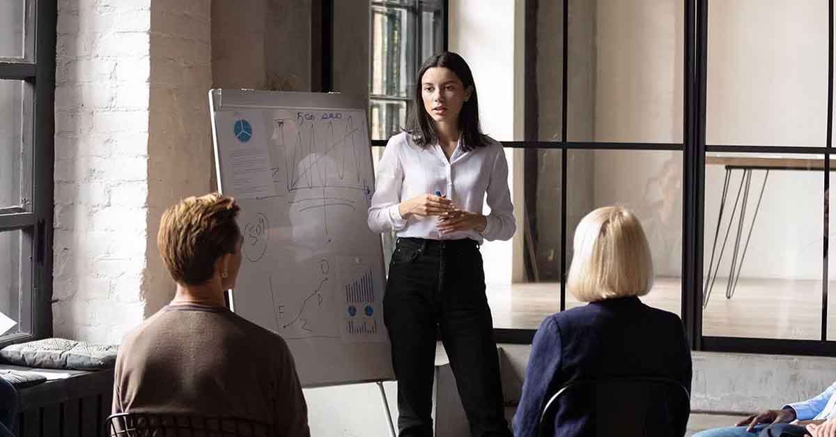 A professional woman giving a business presentation with charts on a whiteboard in a modern office setting.