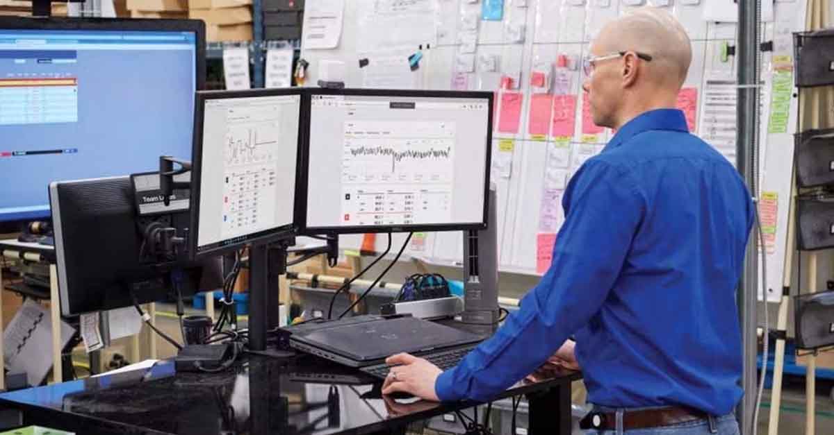 A man monitoring data analytics on dual computer screens in an industrial workspace.