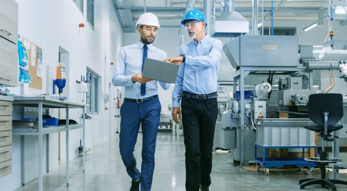 Two engineers wearing safety helmets discussing details on a laptop while walking through a manufacturing facility