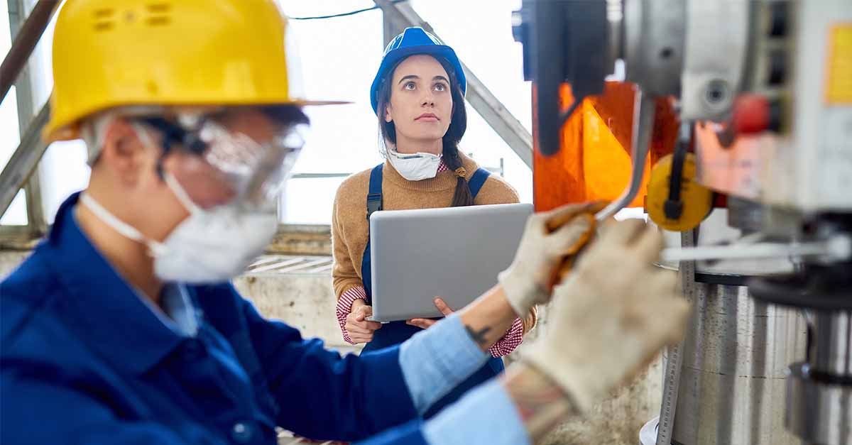 Female engineer in a blue hard hat holding a laptop while observing a worker operating machinery.