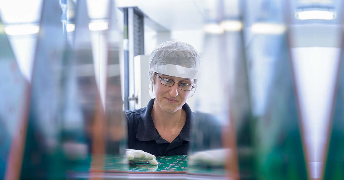 A worker in a lab coat, hair net, and glasses examining a circuit board in a clean, industrial environment