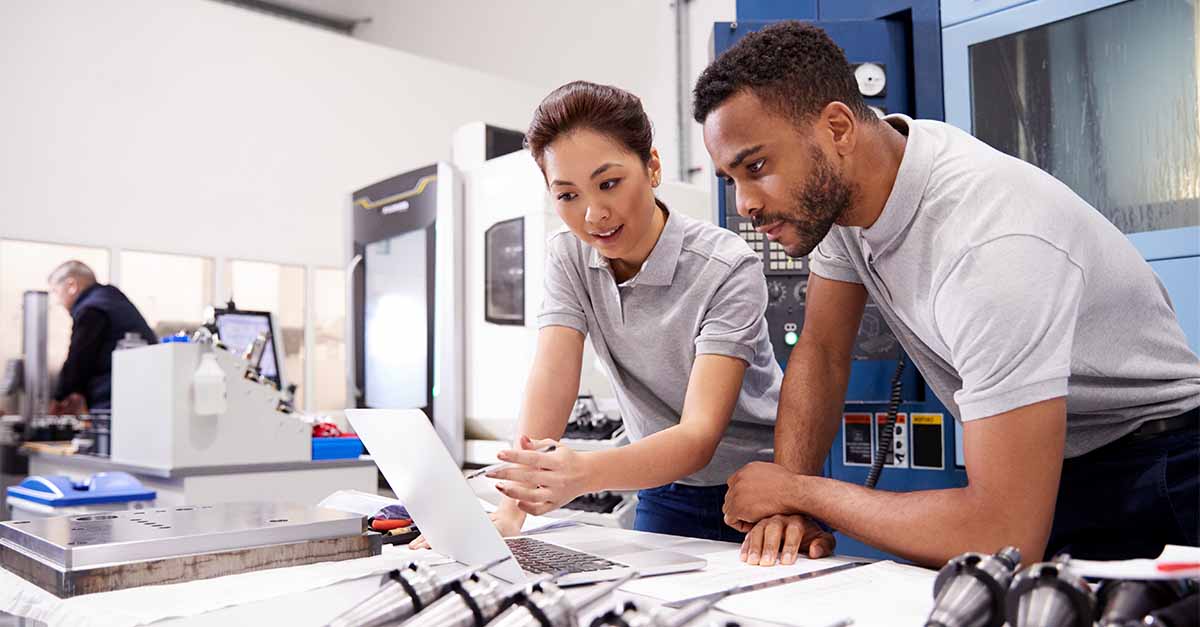 Two engineers in a manufacturing facility reviewing data on a laptop, surrounded by industrial equipment and machinery.