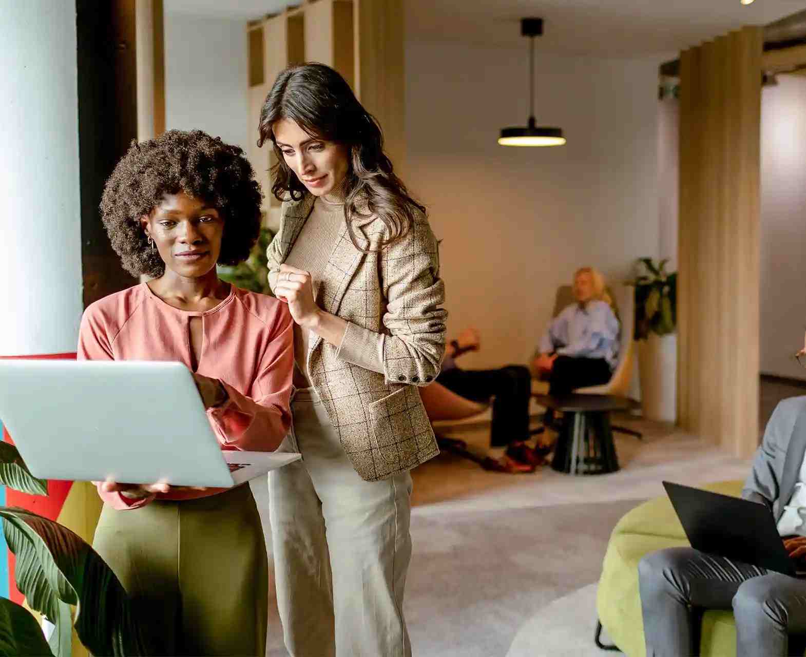 Two professional women collaborating on a laptop in a modern office lounge.