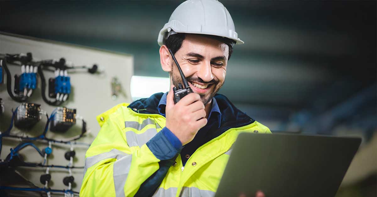 Smiling engineer in a hard hat and high-visibility jacket using a radio while working on a laptop.