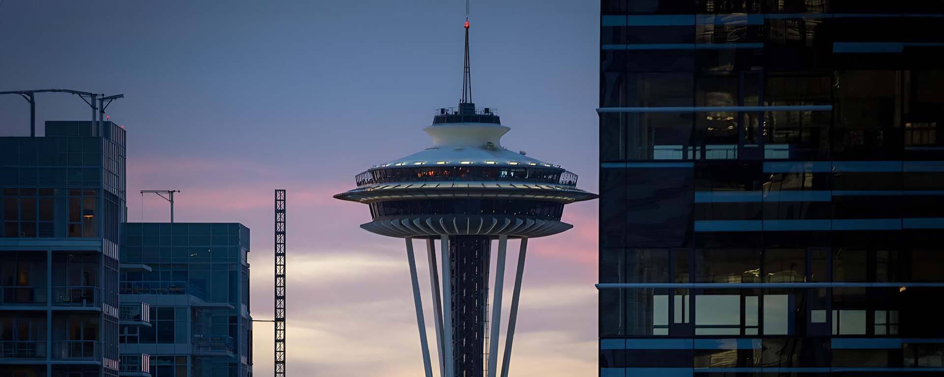 Seattle's Space Needle illuminated at dusk, framed between modern high-rise buildings.