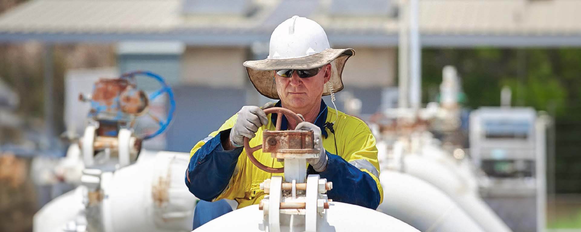 An industrial worker wearing protective gear, including a hard hat, sunglasses, and gloves, operating a valve on a large pipeline system in an outdoor industrial setting.