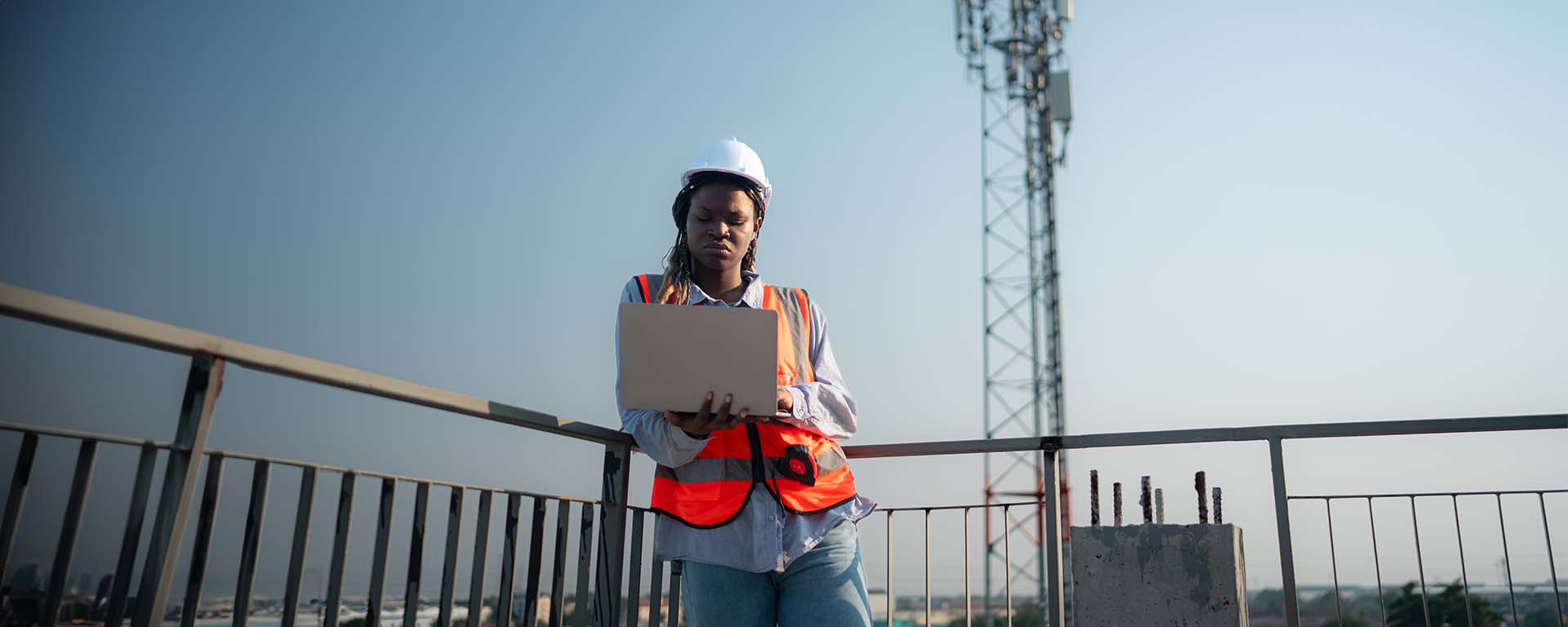 Female construction worker in a safety vest and hard hat using a laptop at an outdoor job site.