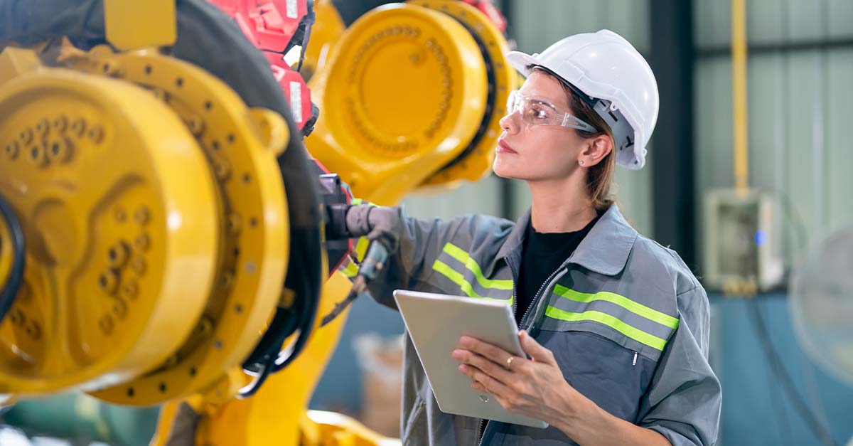 Female engineer in safety gear inspecting industrial machinery while holding a tablet in a manufacturing facility.