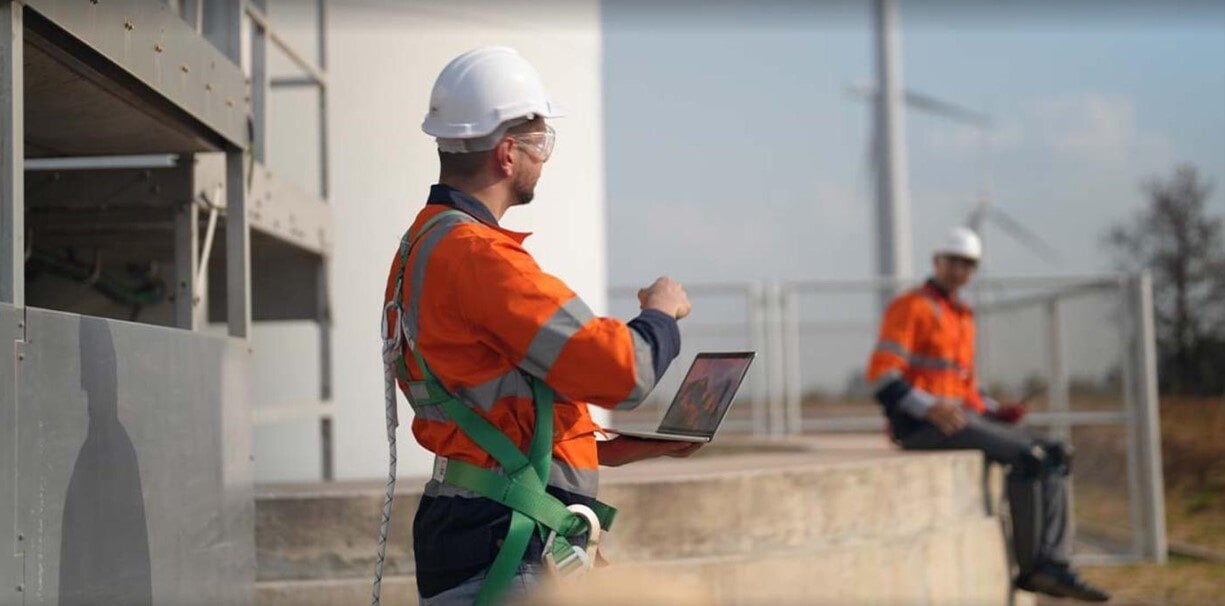 Two construction workers in safety gear at a wind farm site, with one using a laptop while the other sits on a ledge in the background.