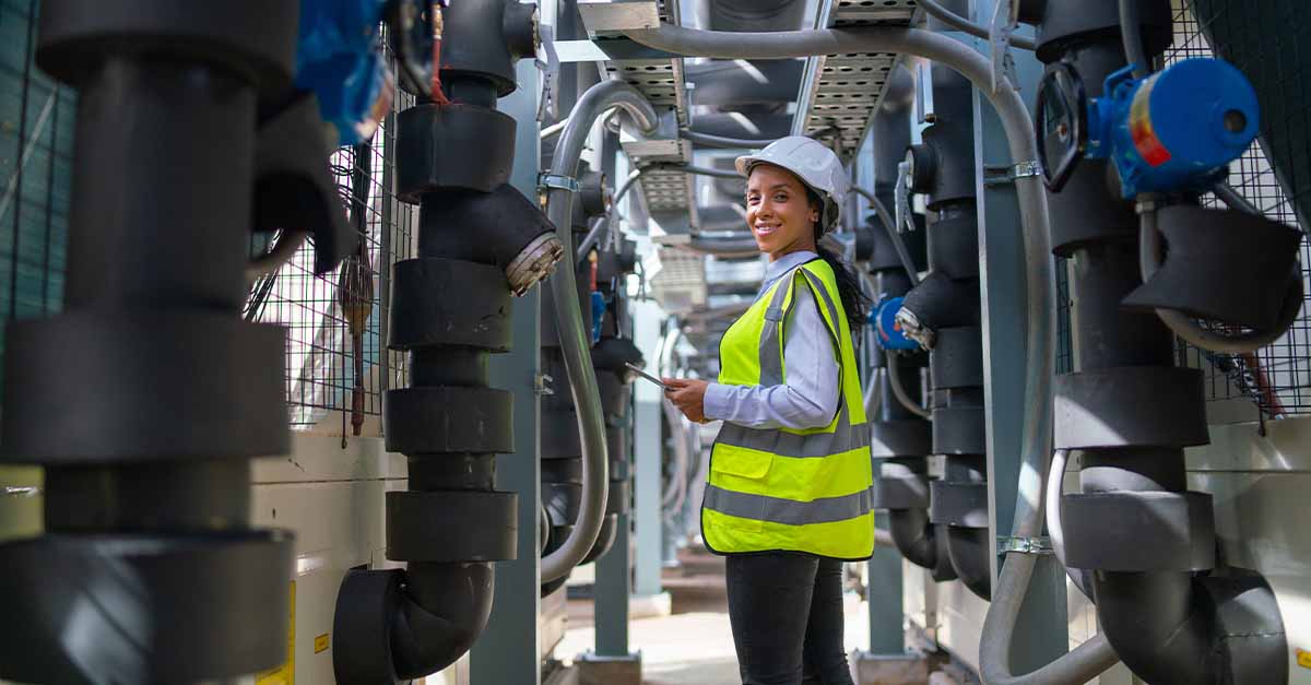 Female engineer in safety gear standing between industrial pipes, holding a tablet.