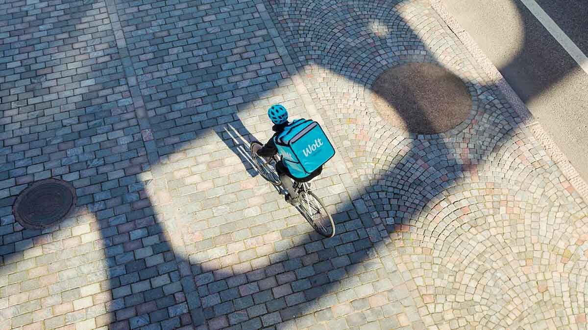 Aerial view of a food delivery cyclist wearing a blue helmet and backpack, riding on a cobblestone street with strong shadows.